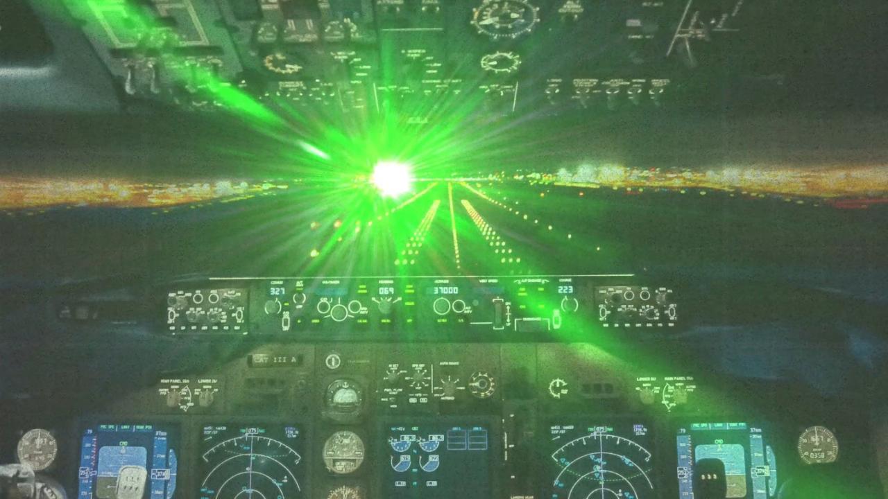 Green laser light floods the windscreen and instrument panel of an airliner cockpit on approach at night, washing out the runway lights ahead